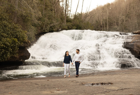A couple holds hands and walks in front of a waterfall.