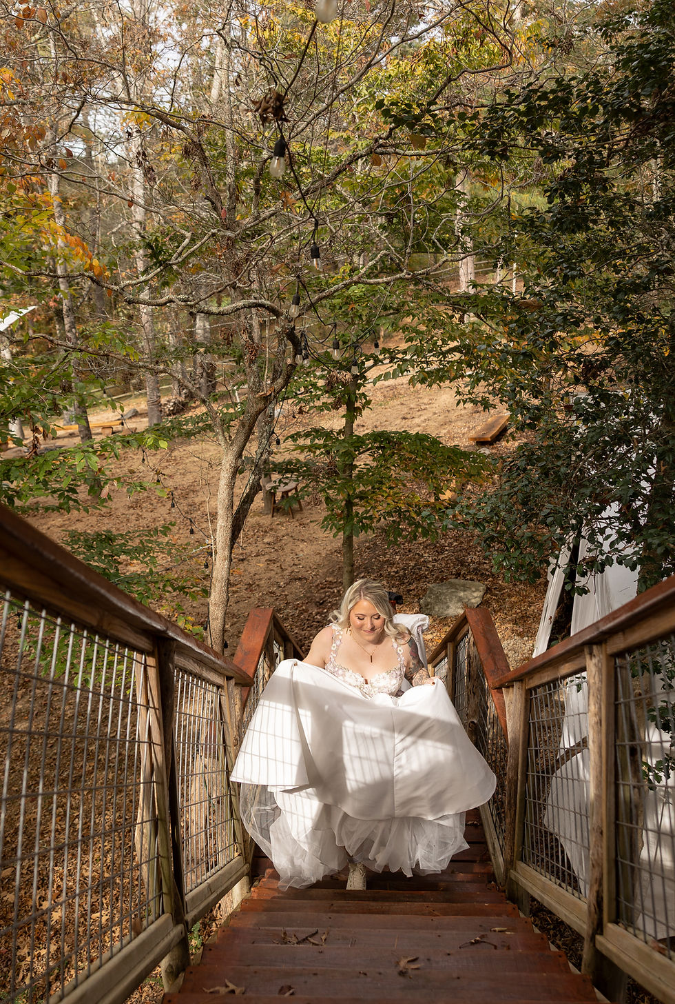 Bride in a white dress ascends wooden stairs outdoors, surrounded by autumn trees and soft sunlight, exuding joy and serenity.