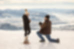 A man proposes to his girlfriend at the top of Jackson Hole Mountain Resort during the winter. They are bundled up in winter clothes as he takes a knee and she smiles. The mountain landscape is vast and covered in snow.