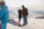 A woman who was just proposed to is still trying to get process everything as she puts her hands on her head and smiles at her fiance. A skier walks by in the foreground.