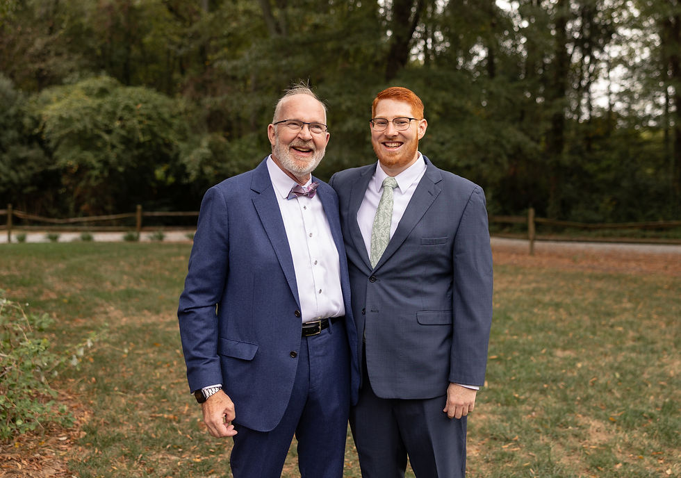 Two men in suits smile in a park with trees in the background. One wears a bow tie, the other a green tie. Mood is joyful.