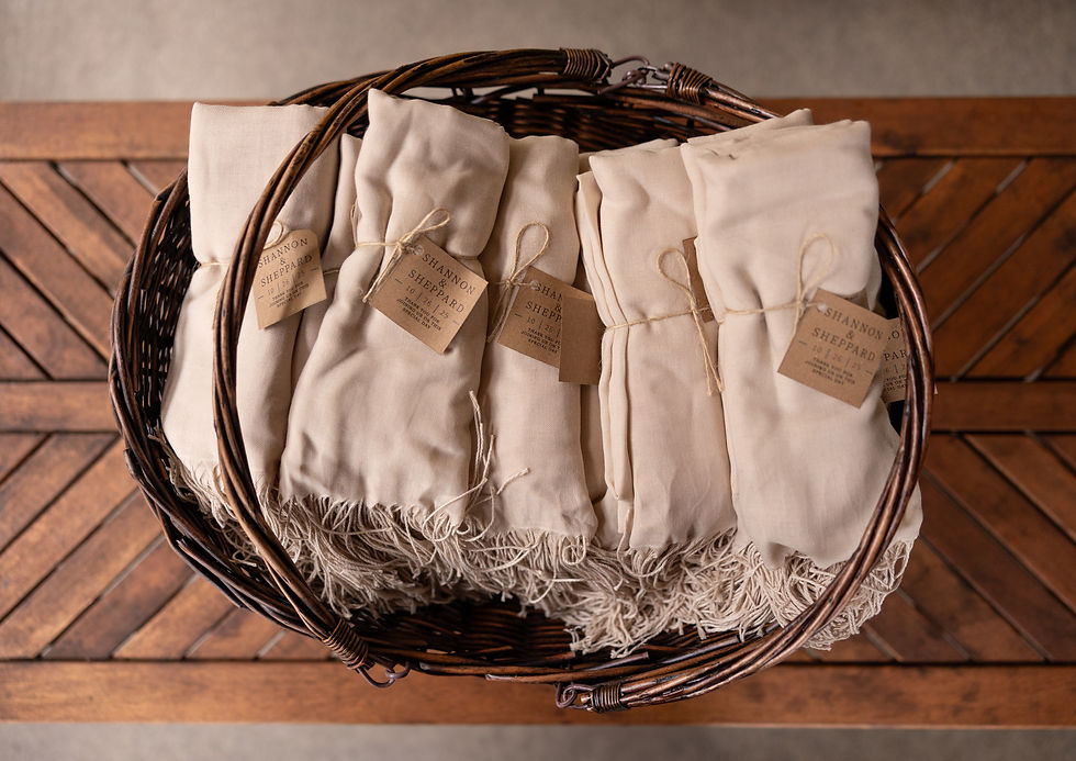 Wicker basket holds rolled beige fabric wraps with twine and tags labeled "Shannon Sheppard." Placed on a wooden patterned surface.