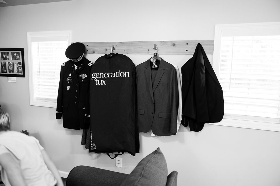 Military uniform, "generation tux" bag, and suits hang on a wall rack. A person is partially visible in the cozy room setting. Black and white.
