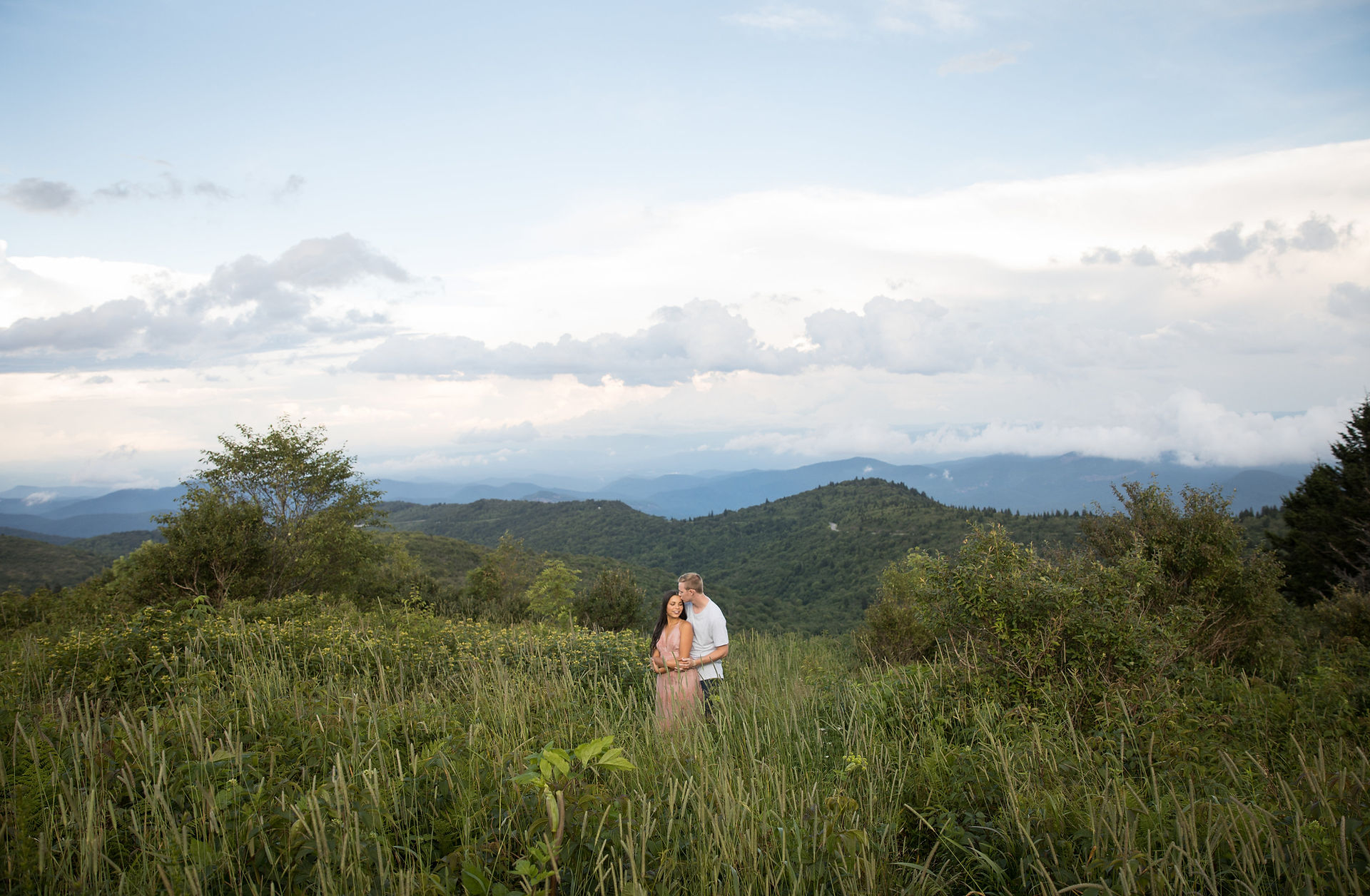 man kissing his fiance on the temple in an open field in the mountains 
