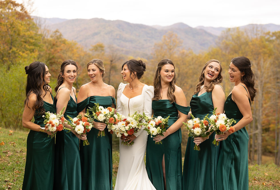 Bride in white and bridesmaids in green dresses holding bouquets, smiling outdoors with autumn trees and mountains in the background.