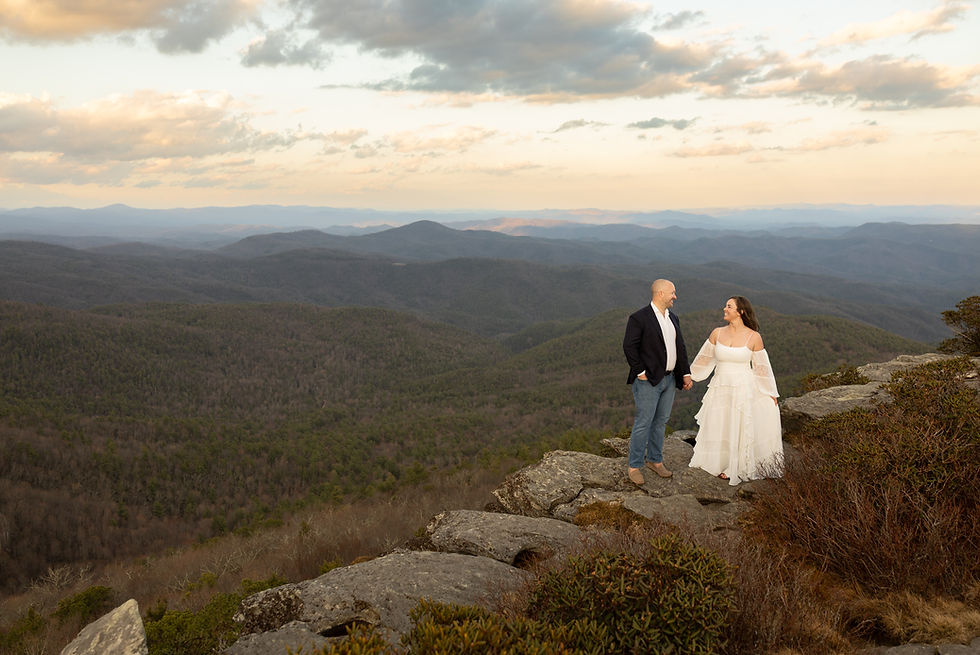 A couple stands on a rocky cliff, holding hands, overlooking green mountains under a cloudy sky. She wears a white dress, he a dark jacket.