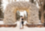 Couple kissing under a large antler arch in snowy park. He wears a cowboy hat, she wears boots. Sign reads "Jackson Hole, WY." Winter trees.
