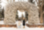 Man kneels proposing to woman under antler arch in snowy park. "Jackson Hole, WY" sign above. Winter coats and boots, trees surround.
