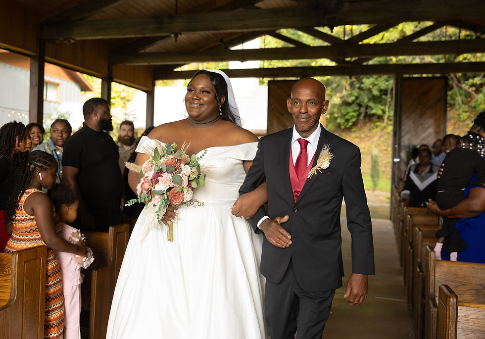 Bride in white gown holding bouquet, walking arm-in-arm with man in suit. Guests watch in wooden chapel with green foliage outside.
