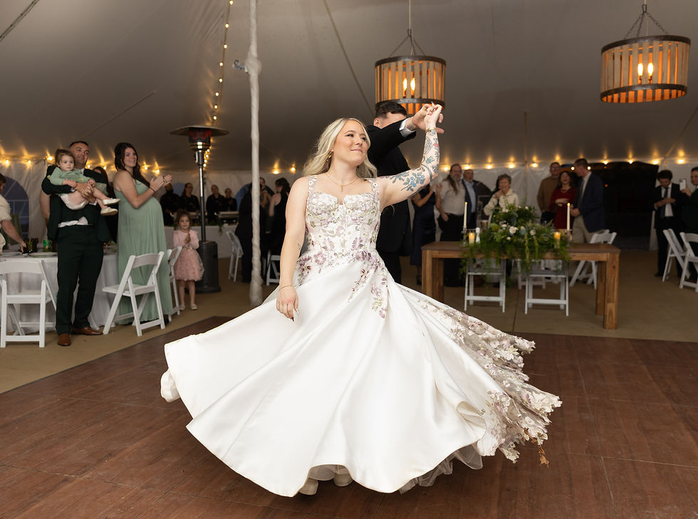 Bride in floral white gown spins on dance floor, groom twirling her. Guests clap in a warmly lit tent with hanging lights and decor.