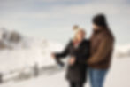 A couple joyfully pops champagne on a snowy mountain, with snow-covered peaks and a fence in the background. They wear winter clothes.