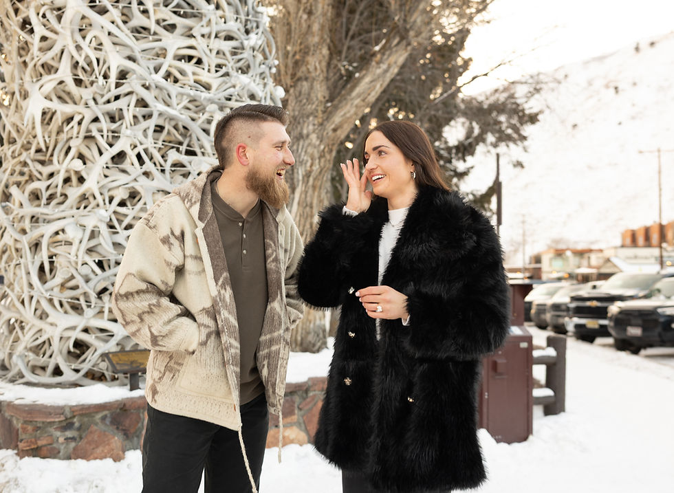 A man and woman in winter coats laugh together outdoors by a large antler sculpture. Snow-covered ground and cars in background.