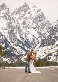 A bride and groom kiss in the middle of a road that looks like it leads right to the mountains in the background that are covered in snow.