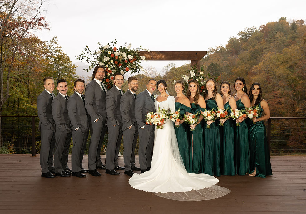 Wedding party outdoors with bridesmaids in green dresses and groomsmen in gray suits. Bride in white holds bouquet; autumn trees in background.