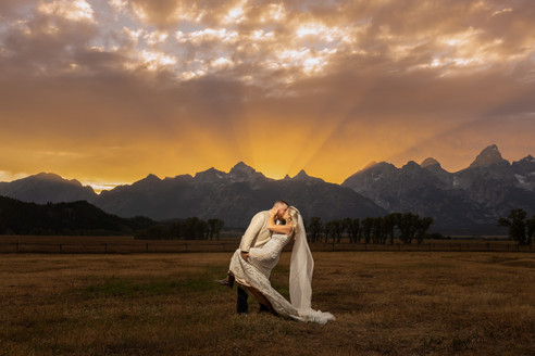 A bride and groom dip kiss during sunset at Mormon Row. The sky is orange and light rays are shooting upward.