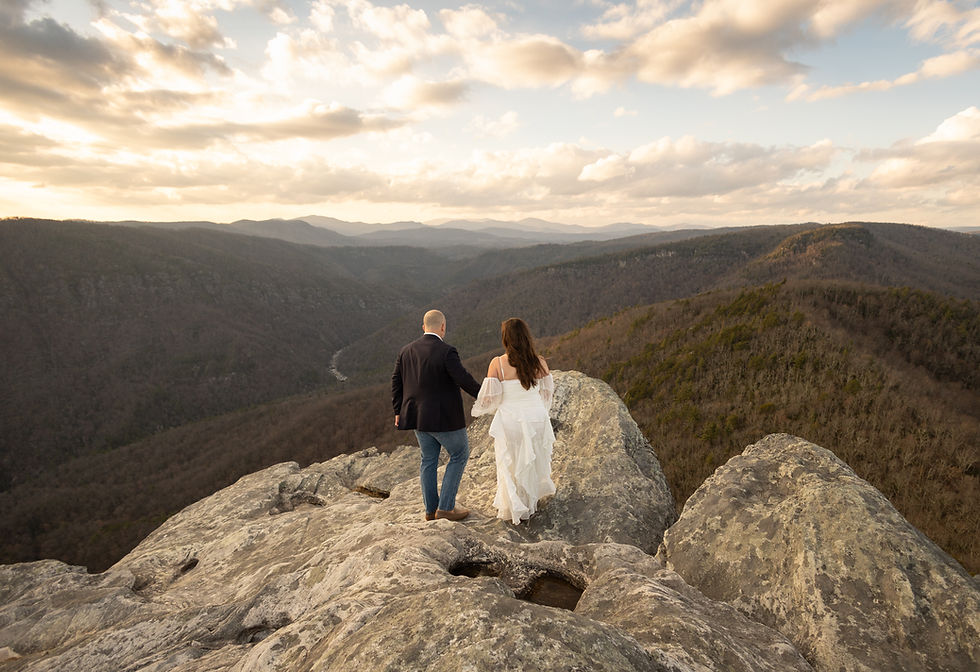 A couple stands on a rocky cliff, holding hands, overlooking expansive mountains. The sky is partly cloudy with a warm, serene atmosphere.