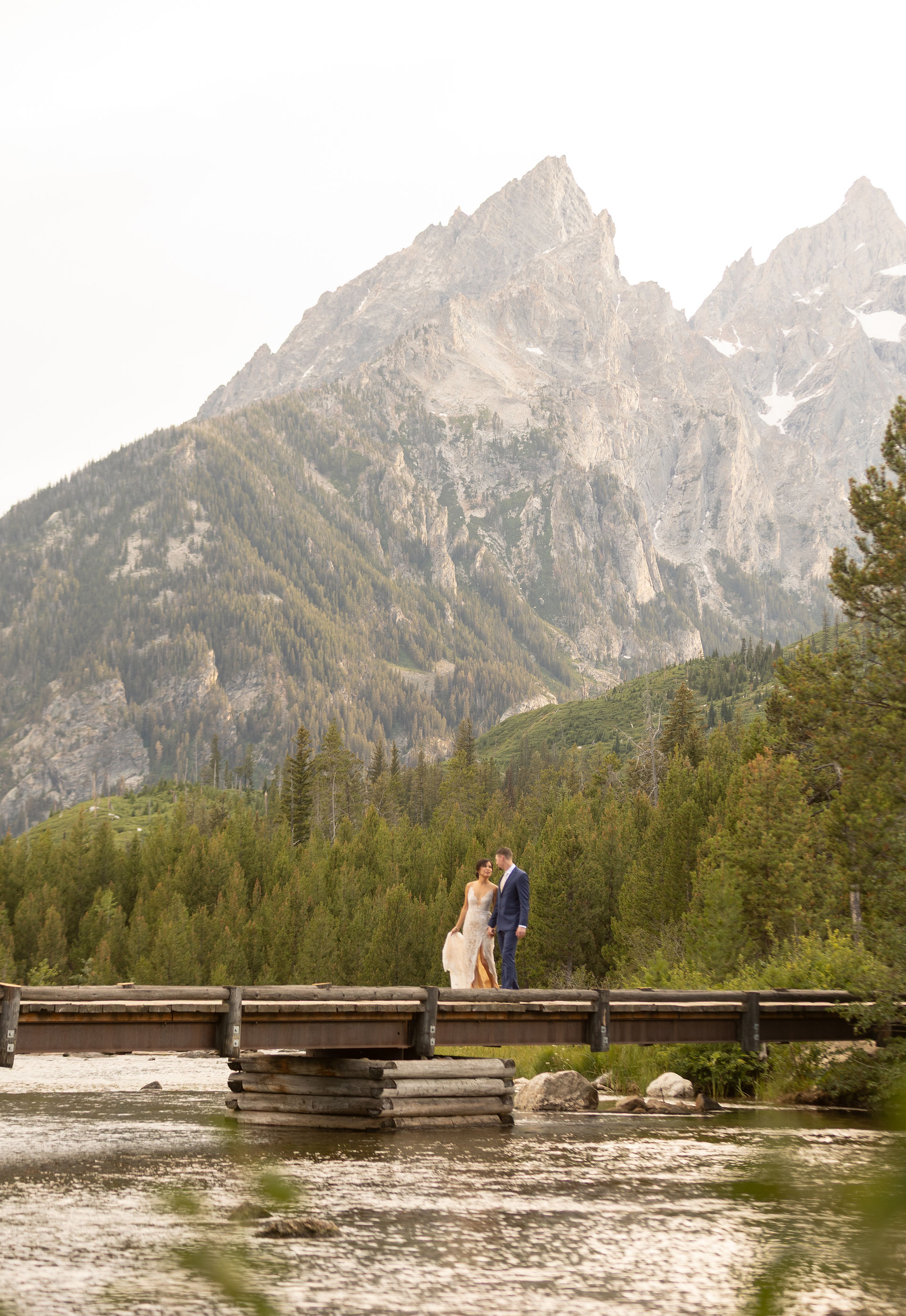 A bride and groom hold hands and walk across a wooden bridge as the mountain tower above them in the background.