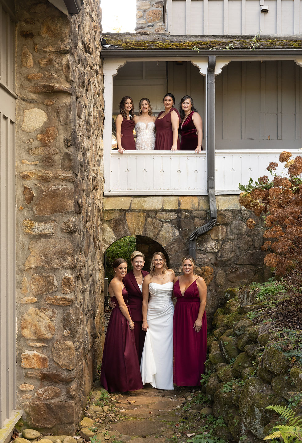 Bridesmaids in burgundy and a bride in white stand in a stone courtyard. Two bridesmaids are on a white balcony above, all smiling warmly.