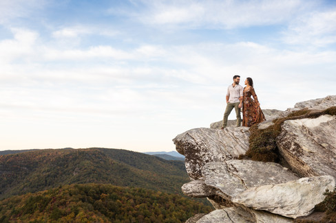 A couple stands on the edge of a cliff while looking at each other with the mountains and a blue sky in the background.  