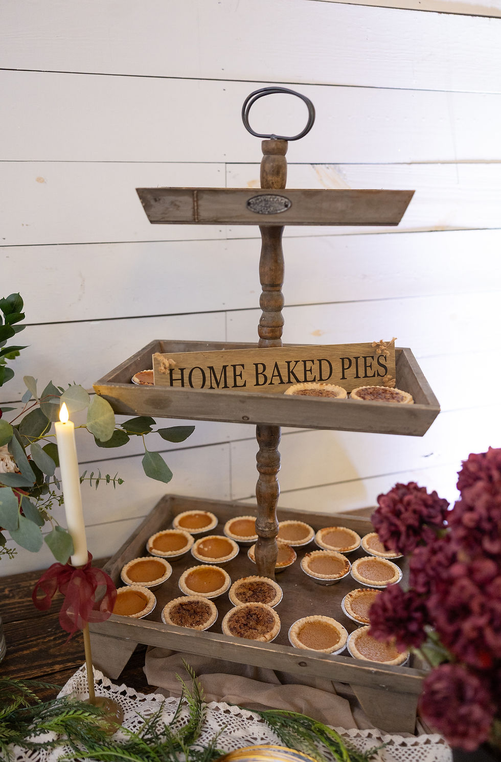 Wooden tray display with assorted pies and "Home Baked Pies" sign. Candle and flowers add a cozy, rustic touch against a white wall.