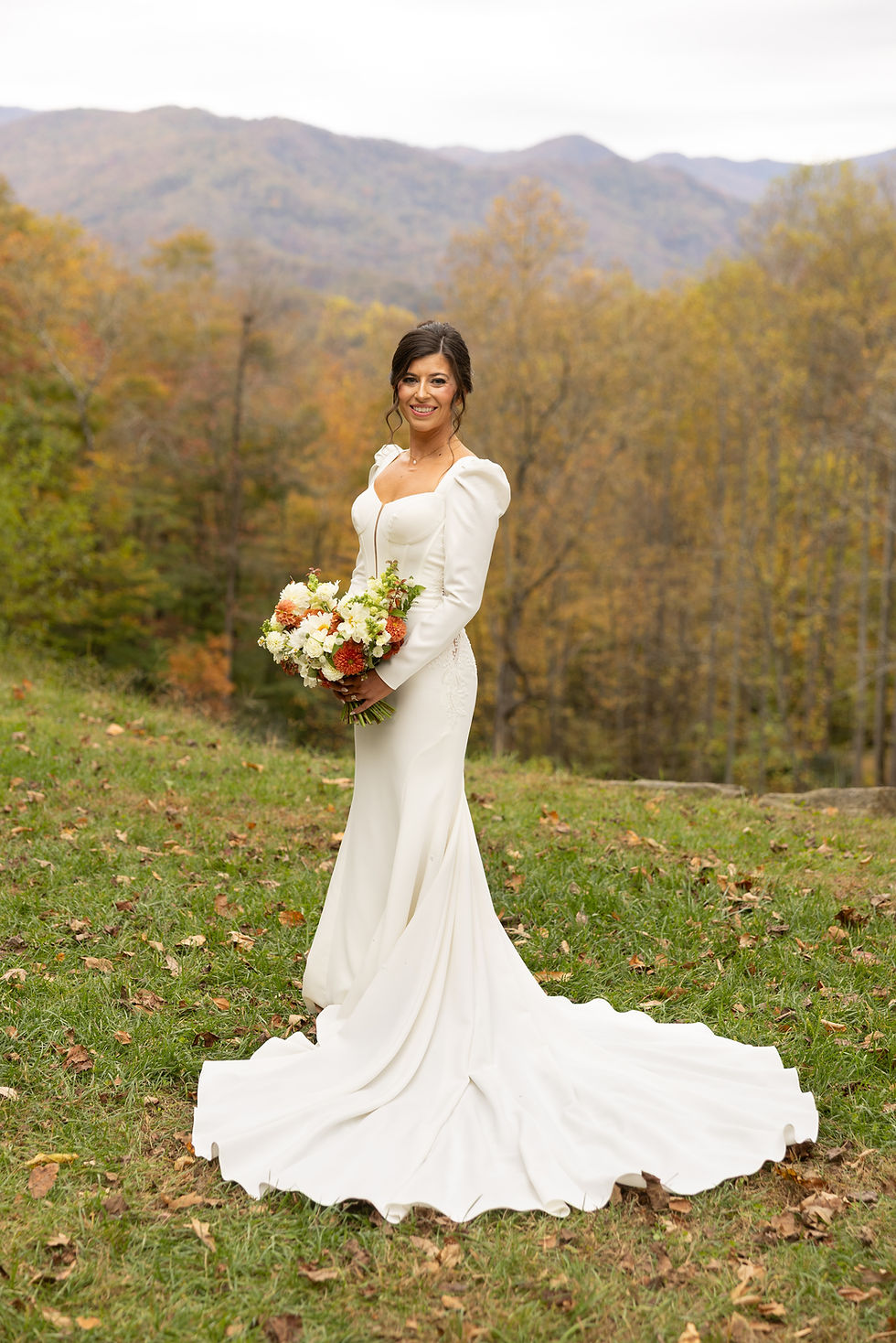 A traditional bridal portrait of a bride smiling at the camera while holding her colorful bouquet. She stands in front of the mountains at the parker mill near Asheville, NC.