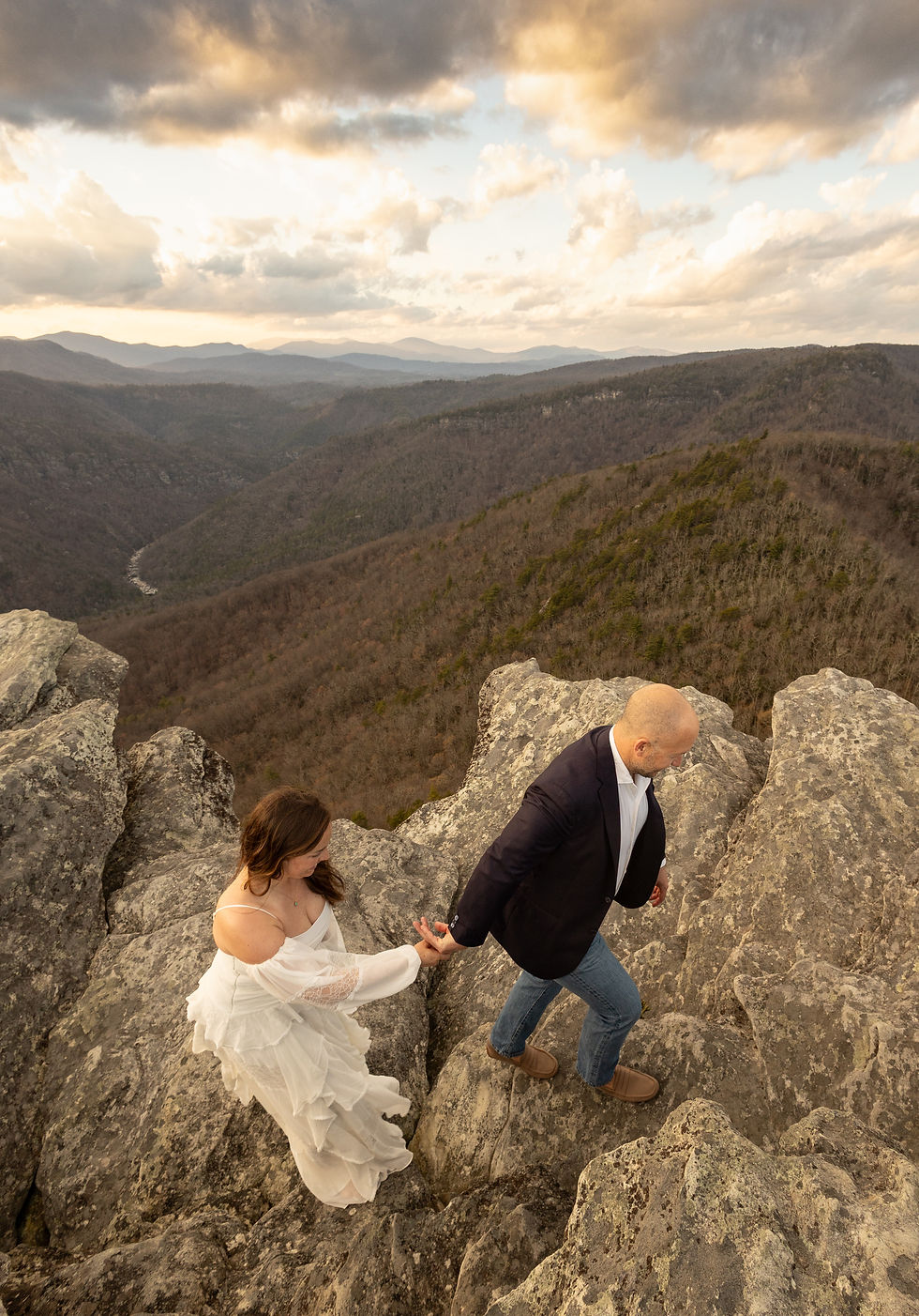 A bride and groom holding hands and hiking across a mountain top at Hawksbill Mountain trail.
