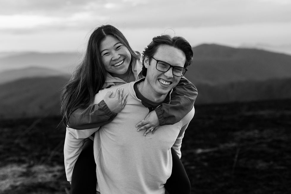 A smiling woman gives a piggyback ride to a man; both are laughing. Mountain landscape in the background. Black and white photo.