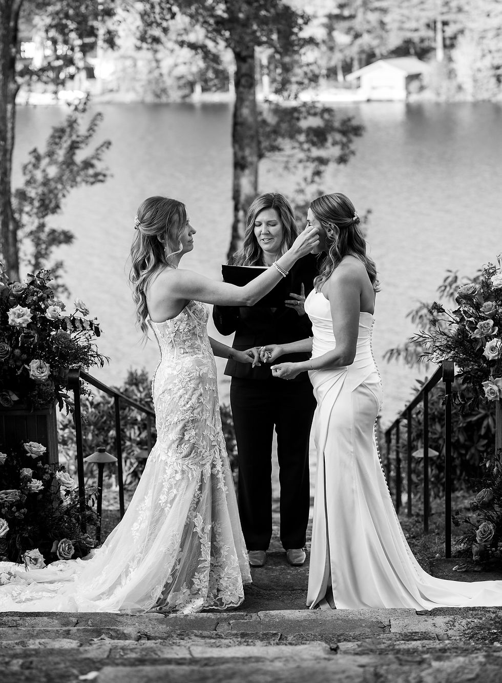 Two brides in elegant gowns hold hands during a lakeside wedding ceremony. A celebrant stands between them. Romantic and serene setting.