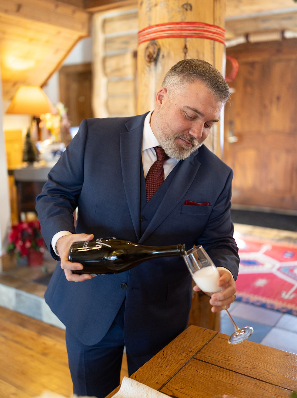 Man in a blue suit pouring champagne into a glass in a rustic, warmly lit room. Wooden table and Christmas decor visible in the background.