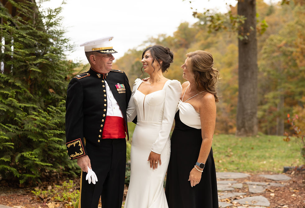Man in military uniform with medals, bride in white gown, and woman in black dress smile together outdoors, surrounded by trees.