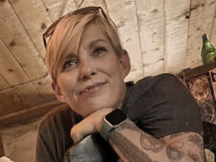 Neurodivergent queer blogger Elle sitting in a rustic cabin, smiling thoughtfully with tattooed arm visible, conveying warmth, authenticity, and lived experience