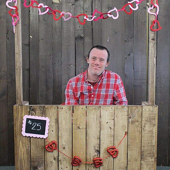 A man with short, dark hair, wearing a red plaid shirt sits behind a Valentine's Day themed booth.