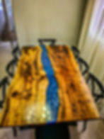 Miniatura: mesa resinada, madeira cor marrom, rio de resina cor azul perolado, cadeiras na cor preta ao redor, cortina clara ao fundo.