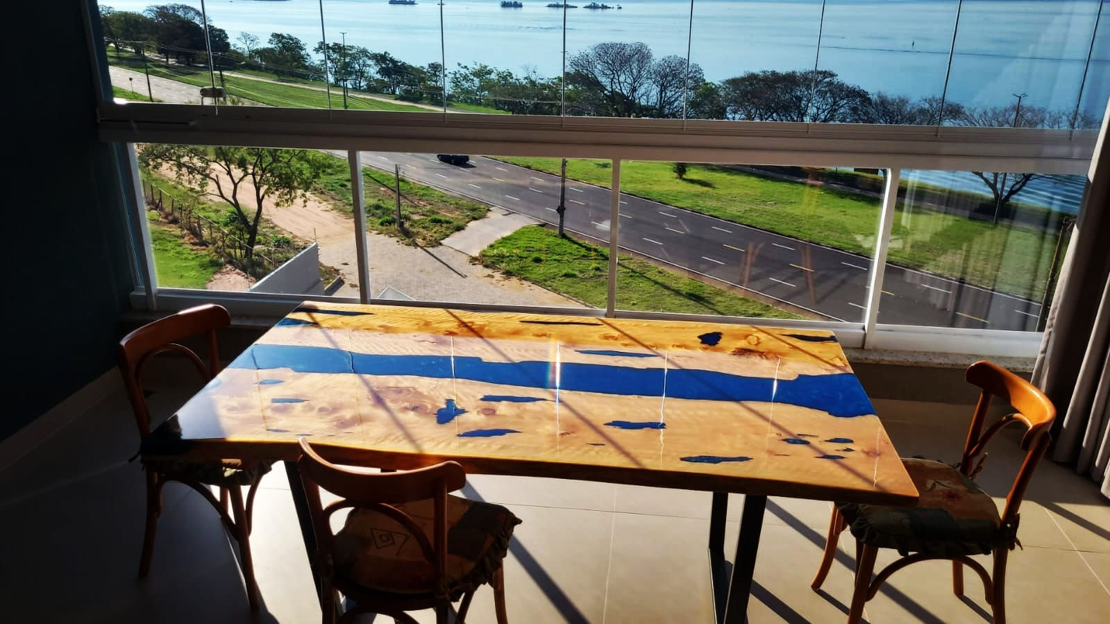 Mesa de jantar madeira clara rio de resina azul no meio, paisagem verde ao fundo