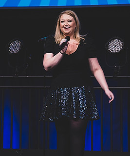 Picture of Kate onstage holding a microphone and presenting the Amazon's Got Talent final. Searing a sparkly skirt.