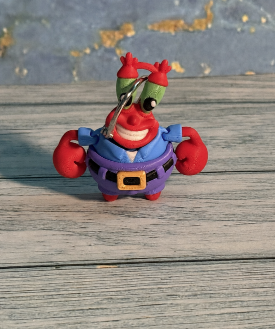 Undersea Money Crab keychain featuring a smiling red crab with tall green eyes, a blue shirt, purple pants