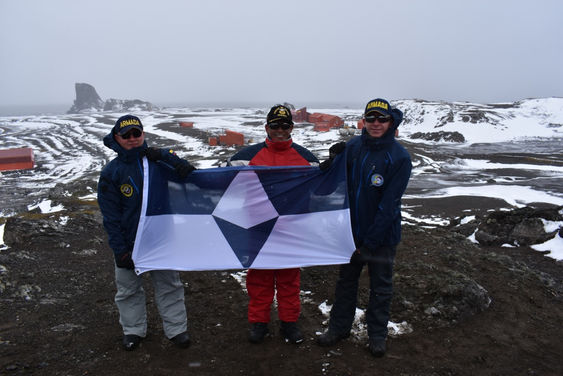 Three people hold the True South flag. Several station buildings are in the background.