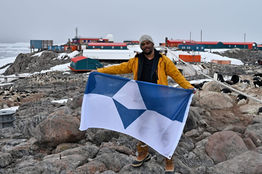 A person holds True South in front of the buildings of Dumont D'Urville Station. 