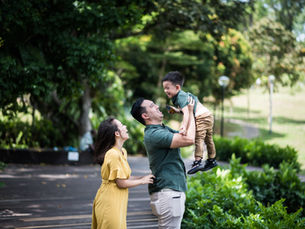 UHNW Smiling parents lift laughing son in a sunny green park, with trees and a path in the background.