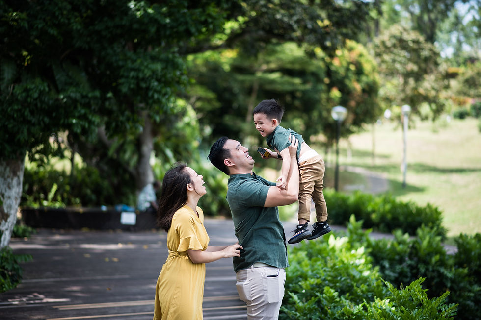 UHNW Smiling parents lift laughing son in a sunny green park, with trees and a path in the background.