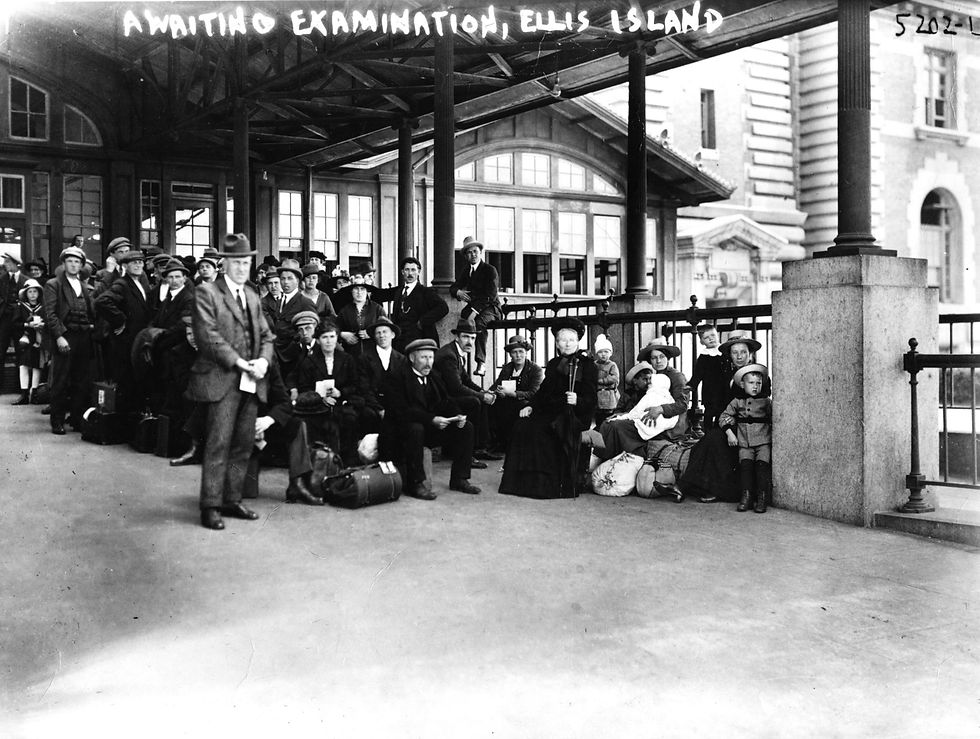 A black-and-white photo of immigrants sitting and standing on the platform at Ellis Island, awaiting examination. They are dressed in early 20th-century clothing, with many carrying luggage. A sign above reads 'Awaiting Examination, Ellis Island.