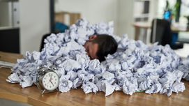 Um homem dormindo em um escritório debaixo de uma pilha de papéis amassados com um relógio à sua frente. | Palavras-chave: RH operacional, RH estratégico, terceirizar rh, bpo de rh, terceirização de rh