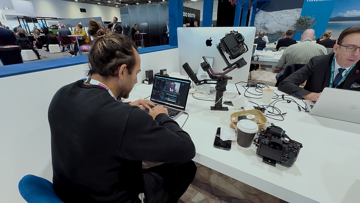 Ben sits at a large white desk in a busy conference centre working at a laptop.