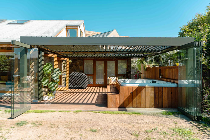 Outdoor entertainment space with a timber deck and hot tub housed under a black louvred pergola. Folding glass doors open the space to the yard, featuring a stone-clad wall, wooden French doors, and a built-in BBQ area.