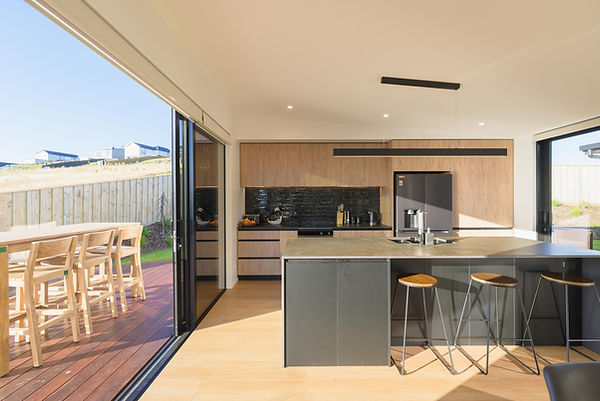 Contemporary kitchen with a large dark grey island and minimalist bar stools, connected by sliding glass doors to a sunny timber deck with outdoor seating. The interior features wood-textured cabinets, a glossy black subway tile backsplash, and a linear black pendant light.