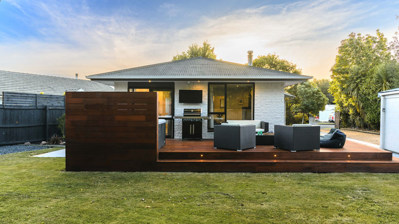 Backyard view of a contemporary white brick home with a large timber entertainment deck at dusk. The deck features integrated step lighting, a dark wood privacy screen, and a modern outdoor lounge set with grey wicker chairs and a built-in BBQ station.
