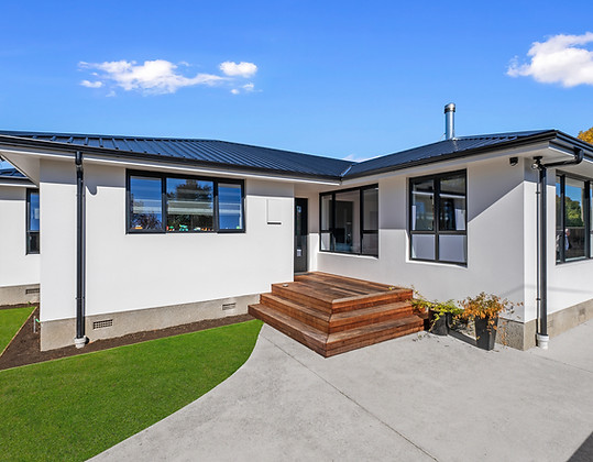 Exterior view of a modern L-shaped single-story home with white stucco walls and a black corrugated metal roof. The design features black-trimmed windows, a wooden entry deck with wrap-around steps, and a clean concrete driveway bordering a manicured green lawn.