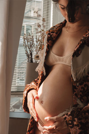 Lifestyle maternity photo of a woman standing in her bathroom, holding her baby bump in natural light