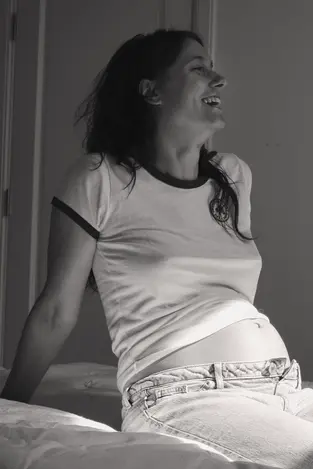 Black and white casual pregnancy photo of a woman sitting on her bed, laughing and showing her baby bump