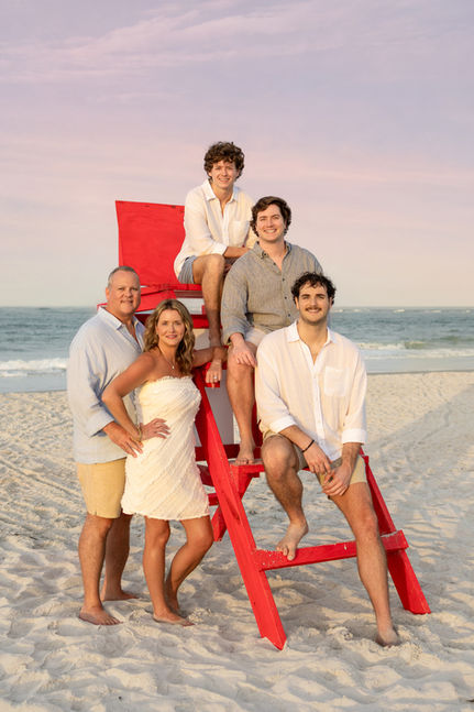 A family of five poses playfully around a red lifeguard chair on the beach at golden hour, with three young adult sons and their parents.